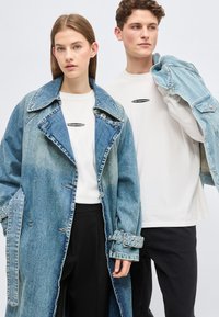 Νεαρή γυναίκα που φοράει μακρύ τζιν παλτό και λευκό πουκάμισο, νεαρός άνδρας που κρατάει τζιν μπουφάν στον ώμο του, φορώντας λευκό πουκάμισο και μαύρο παντελόνι.