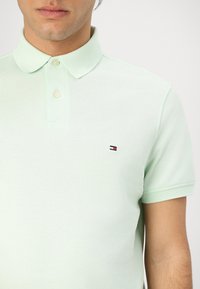 Homme portant un polo vert clair avec deux boutons et petit logo Tommy Hilfiger sur la poitrine gauche.