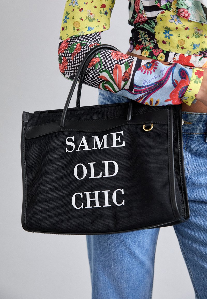 Must mustre kott, millel on valge tekst "SAME OLD CHIC", kahte käepidet ja kuldne aksessuaaride detail.