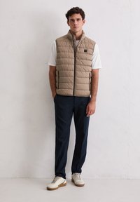 Beige gesteppte Weste mit hohem Kragen und Frontreißverschluss, getragen über einem weißen Hemd. Kombiniert mit marineblauen Hosen und hellen Sneakern. Einfaches und funktionales Design.