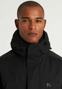 Juodas švarkas su aukštu apykakle, reguliuojamomis raištelėmis ir maža logotipo emblema kairėje krūtinės pusėje. Lygus audinio tekstūra, minimalus dizainas.