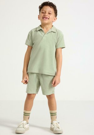 Smilende ung dreng iført lysegrøn polo og shorts, beige sokker med grønne striber og hvide sneakers, stående på ensfarvet baggrund.