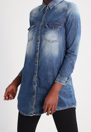 Bluzka z długim rękawem z wyblakłego niebieskiego denimu z zapięciem na guziki z przodu i dwoma kieszeniami na piersi, noszona na czarnych spodniach na stojącej osobie.