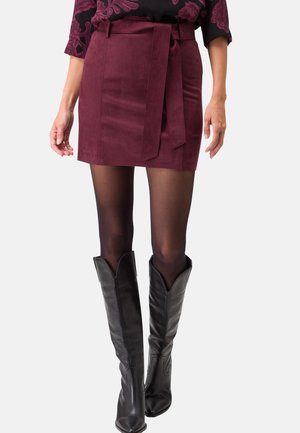 Femme portant une mini-jupe en daim bordeaux avec une ceinture à nouer, des collants noirs transparents et des bottes en cuir noires montant jusqu'aux genoux.