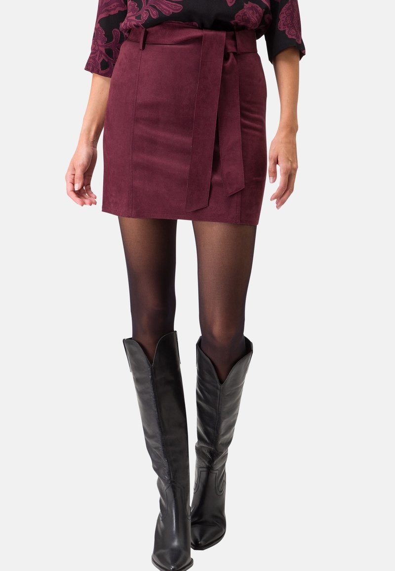 Femme portant une mini-jupe en daim bordeaux avec une ceinture à nouer, des collants noirs transparents et des bottes en cuir noires montant jusqu'aux genoux.
