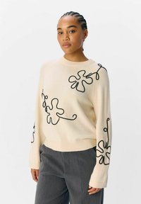 Maglione crema con ricami floreali neri, colletto e polsini a costine, vestibilità comoda, abbinato a pantaloni grigi a trama.