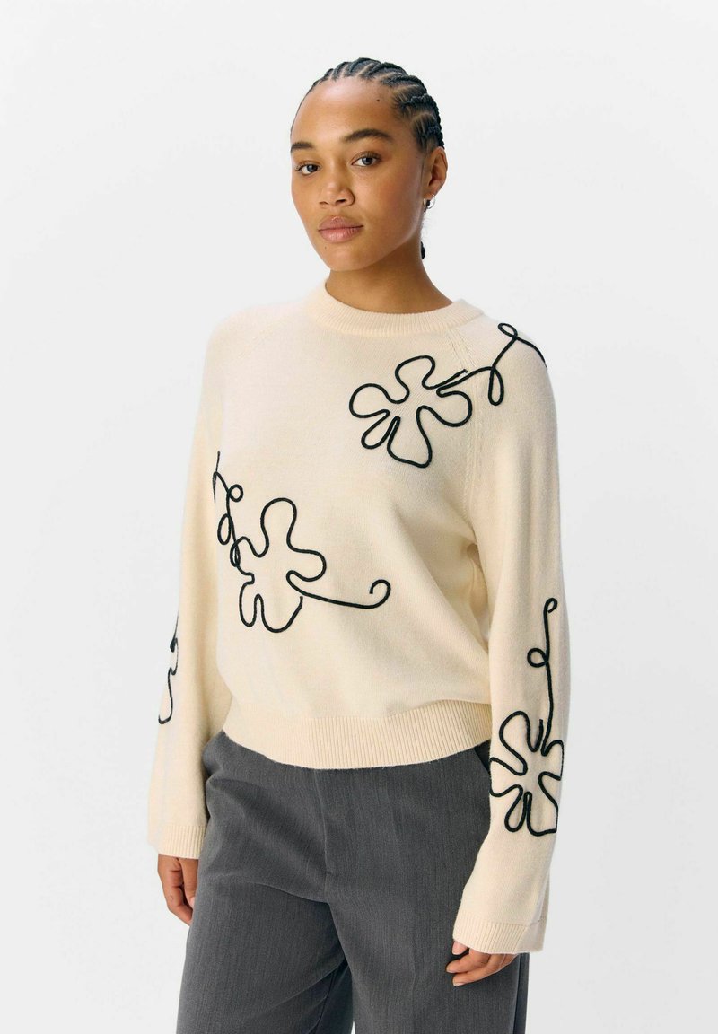 Pull crème avec broderie florale noire, col et poignets côtelés, coupe décontractée, associé à un pantalon texturé gris.