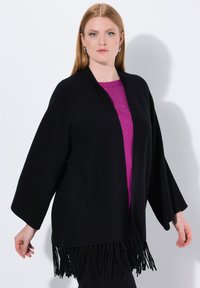 Schwarzer Poncho mit weiten Ärmeln und fransigem Saum, über einem lila Top getragen. Glatte Textur, offenes Design, elegante Silhouette.