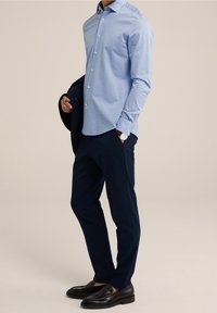 Camisa de botones azul claro de manga larga, combinada con pantalones ajustados azul marino y mocasines negros, sosteniendo un blazer oscuro sobre el brazo.