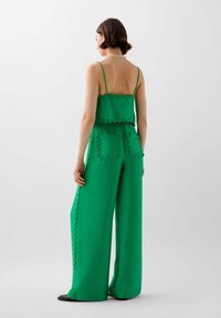 Zelený dvoudílný outfit se skládá z krčeného topu s vroubkovanými okraji a kalhot s širokými nohavicemi s vroubkovanými kapsami. Hladká textura.