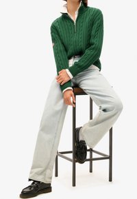 Vihreä neulottu villapaita palmikkokuvioilla ja vetoketjulla kaulassa, yhdistetty vaaleansinisien farkkujen ja mustien loafersien kanssa. Malli istuu metallijakkaralla.