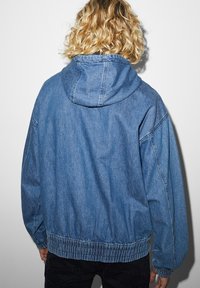 Denimjacka med huva i ljusblått, med rynkad nederkant och ärmar. Tyget har en slät yta med minimala sömdetaljer.