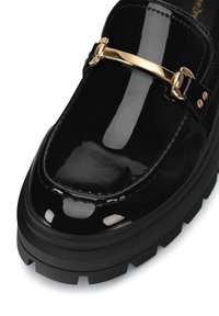 Must valge nahast loafer, millel on paks must tald, kuldne baar aktsent rihmal ja dekoratiivne tikand äärtes.