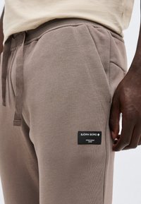 Κοντινό πλάνο στα καφέ παντελόνια jogger με ελαστική μέση και κορδόνι, με ετικέτα "BJÖRN BORG Stockholm 1956" στο μηρό, φορεμένα από άτομο με σκούρο δέρμα.