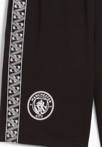 Svarta shorts med en vit logotyp av Manchester City tillsammans med en skeppsgrafik. Sidoflik har ett geometriskt mönster i vitt. Slät textur.