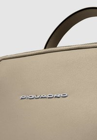 Bolso de piel beige con textura suave, que presenta un logo plateado. Diseño minimalista con asa superior y detalles de costura sutiles.