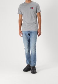 Camiseta de algodón gris con un pequeño bolsillo con logo multicolor, combinada con vaqueros de mezclilla azul claro y zapatillas negras con estampado.