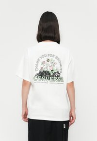 Persona con cabello oscuro de longitud hasta los hombros que lleva una camiseta blanca de Converse con un diseño de flores y abejas y el texto "Gracias por crecer."