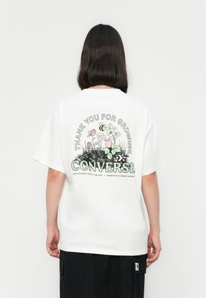 Personne avec des cheveux noirs mi-longs portant un T-shirt blanc Converse avec un design floral et d'abeille et le texte "Merci d'avoir grandi."