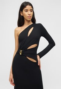 Γυναίκα που φοράει μαύρο φόρεμα με έναν ώμο, με κοψίματα στο στήθος και στο πλάι, με χρυσό διακοσμητικό κούμπωμα στη μέση.