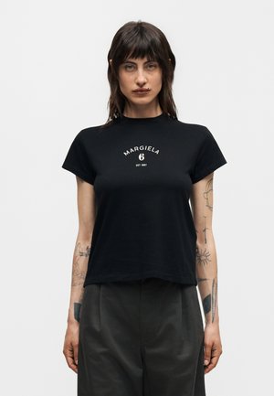 Γυναίκα με σκούρα μαλλιά, φορώντας μαύρο T-shirt Margiela 6 και σκούρο παντελόνι, εμφανίζοντας τατουάζ και στα δύο αντιβράχια σε απλό φόντο.