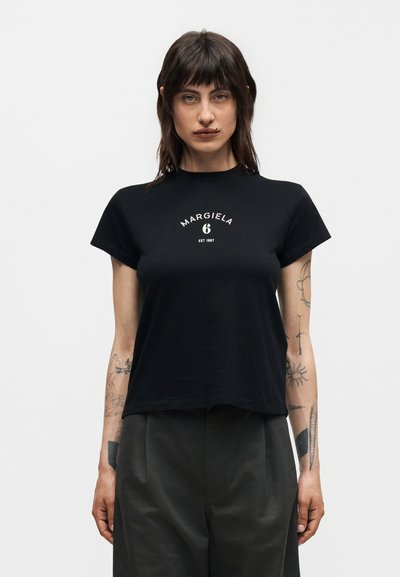 Γυναίκα με σκούρα μαλλιά, φορώντας μαύρο T-shirt Margiela 6 και σκούρο παντελόνι, εμφανίζοντας τατουάζ και στα δύο αντιβράχια σε απλό φόντο.
