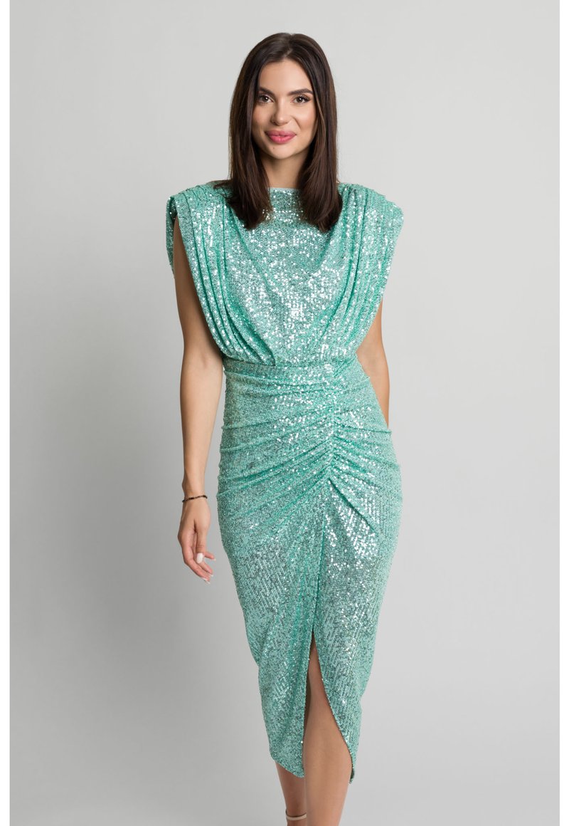 Kobieta w błyszczącej, bezrękawnikowej turkusowej sukience z cekinami, z poduszkami na ramionach, marszczonym pasem i rozcięciem z przodu, stojąca na szarym tle.