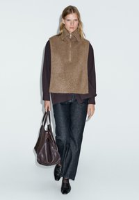 Top marrone senza maniche con colletto alto e zip, sovrapposto a una maglietta a maniche lunghe scura, abbinato a jeans in denim scuro e una grande borsa marrone.