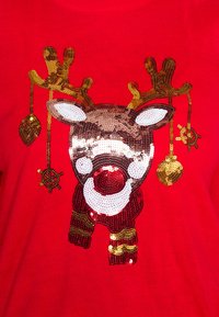 Volto di renna decorato con paillettes, naso rosso e ornamenti d'oro appesi alle corna su uno sfondo di tessuto rosso.