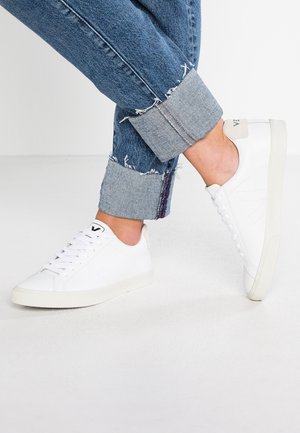 Persona con vaqueros azules vueltas y zapatillas blancas bajas de pie sobre un suelo blanco con un pie ligeramente levantado.