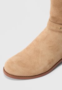 Vaaleanruskeat mokkanahkaiset nilkkurit, joissa pyöreä kärki, sileä pinta ja ruskea kumipohja. Varustettu remmillä, jossa pieni kullanvärinen niittikoriste.