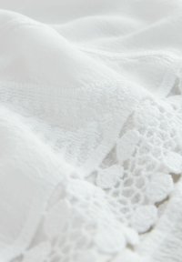 Tissu blanc avec une surface texturée, doté d'une délicate bordure en dentelle aux motifs complexes. Texture douce et lisse, mettant en valeur les détails fins.