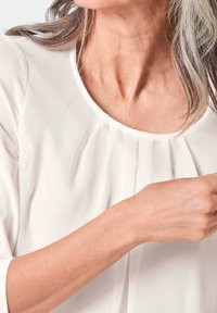 Weiße Bluse mit rundem Ausschnitt, vorne mit Falten, Dreiviertelärmeln und glatter Stofftextur.