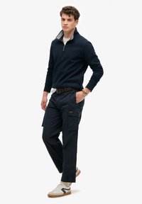 Marineblå pullover med lynlås krave over en hvid skjorte, parret med mørke cargobukser. Har et bælte og hvide sneakers med sorte accenter.