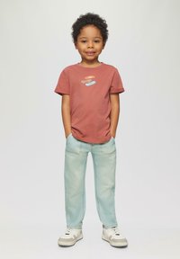 Camiseta de color óxido con diseño gráfico, elementos en azul claro y amarillo. Pantalones azul claro con una textura desgastada. Zapatillas blancas con detalles en gris.