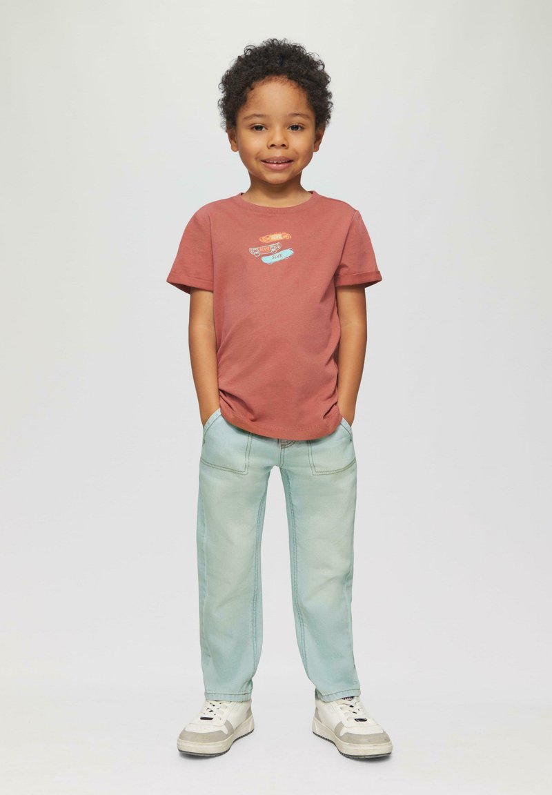 Camiseta de color óxido con diseño gráfico, elementos en azul claro y amarillo. Pantalones azul claro con una textura desgastada. Zapatillas blancas con detalles en gris.