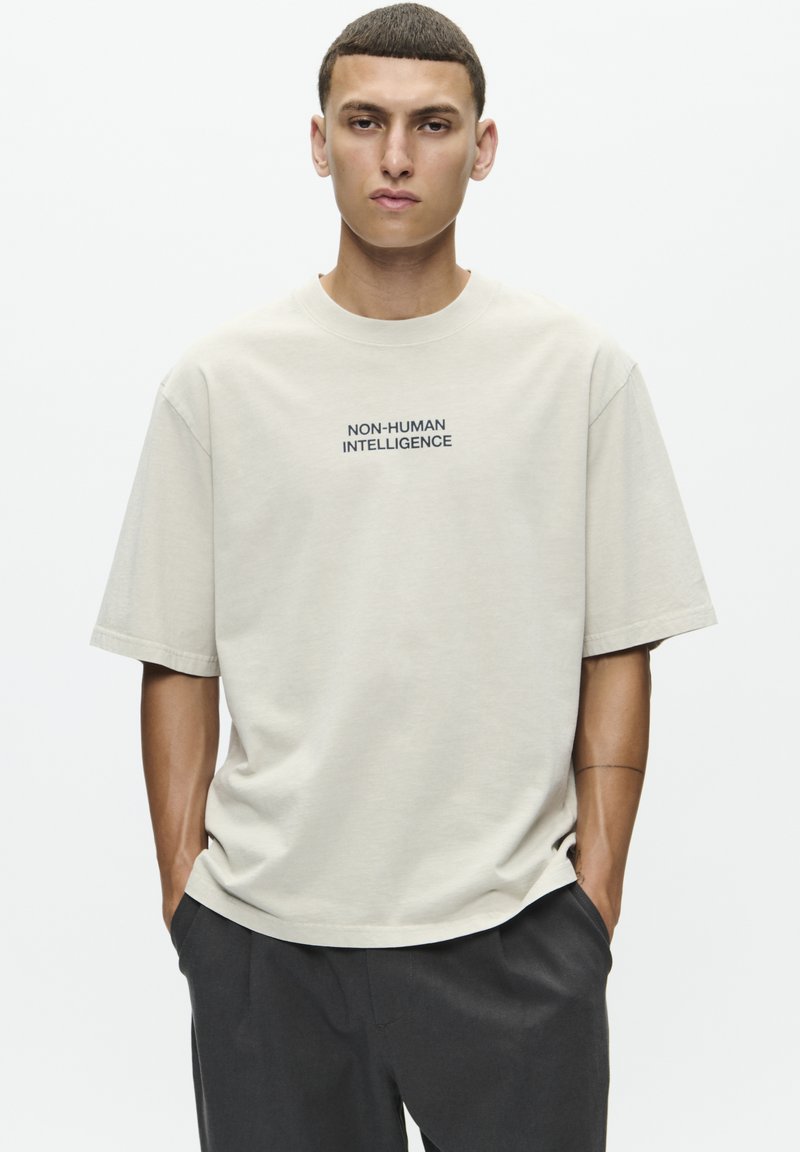 T-shirt en coton beige avec des manches courtes, un col rond, portant l'inscription "NON-HUMAN INTELLIGENCE" imprimée en noir sur la poitrine.