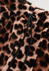 Pantaloni tapered cu imprimeu de leopard dintr-un material moale de catifea, cu detalii de prindere neagră și o textură subtilă de rigle verticale în tonuri de maro și roz.