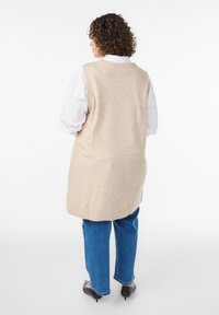 Žena s kučeravými vlasmi, oblečená v béžovej dlhej veste cez bielu košeľu, modré džínsy a hnedé lodičky, stojaca chrbtom k pozorovateľovi na bielom pozadí.