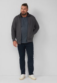 Giacca zip grigia con cappuccio, indossata sopra una maglietta blu, abbinata a jeans scuri e sneaker bianche. Il modello sta in piedi con le mani nelle tasche.
