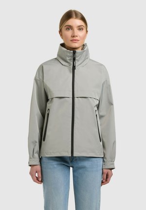 Nő világosszürke, magas nyakú dzsekiben fekete cipzárral és oldalzsebekkel, világoskék farmernadrágot viselve, előre nézve.