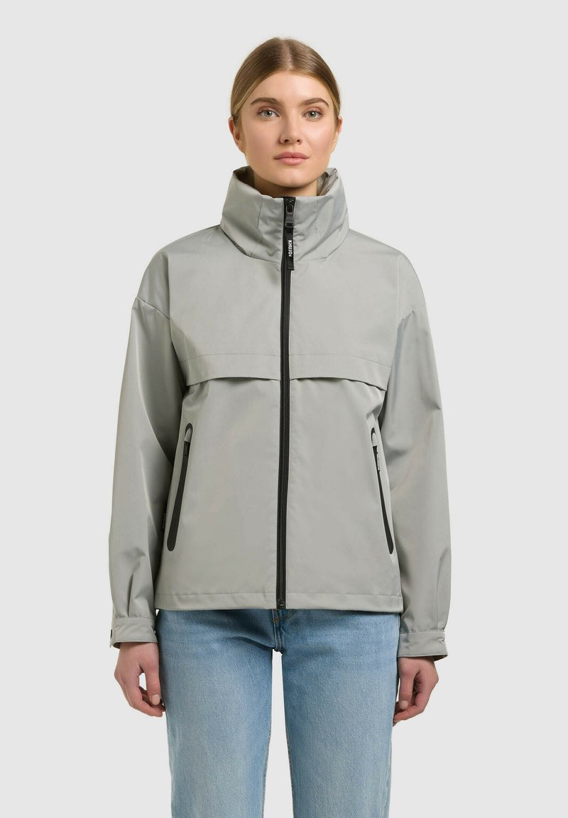 Femme portant une veste gris clair à col montant avec une fermeture éclair noire et des poches latérales, associée à un jean bleu clair, face à l'avant.