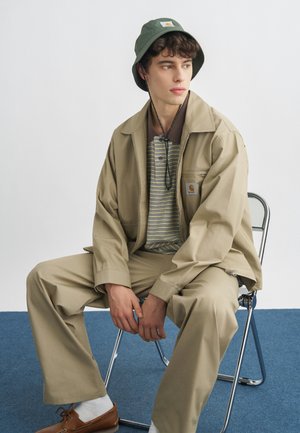 Młody mężczyzna siedzący na metalowym krześle, ubrany w beżową kurtkę i spodnie, pasiastą koszulę, oliwkowozielony kapelusz bucket, białe skarpetki i brązowe buty.