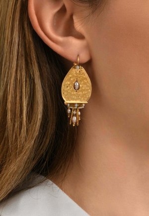 Boucle d’oreille dorée en forme de goutte avec une perle centrale et des perles suspendues ainsi que des chaînes en or, portée à l’oreille d’une femme aux cheveux châtain clair.