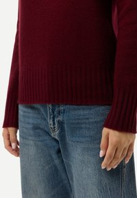 Personne portant un pull côtelé bordeaux et un jean en denim bleu, les mains détendues le long du corps.