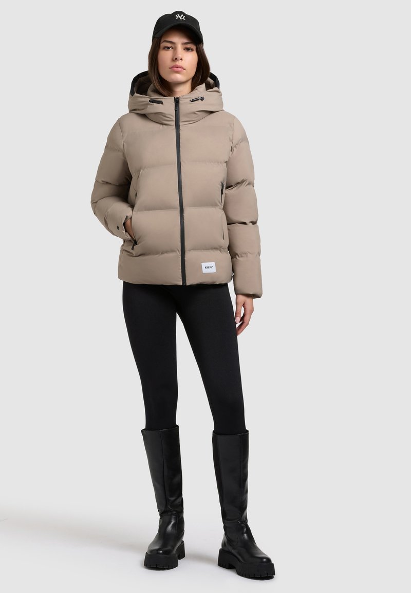 Chaqueta de plumas beige con capucha, cremallera negra y parche de logo. Combinada con leggings negros y botas altas negras. Textura suave y acolchada.