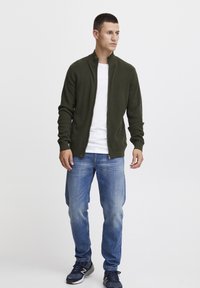 Oliven grønn zip-up genser med ribbet struktur, båret over en hvit t-skjorte. Kombinert med lyseblå jeans og mørke sneakers.
