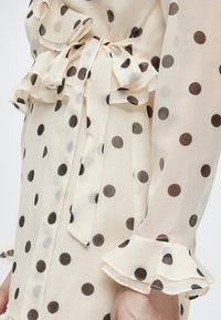 Φόρεμα από κρεμ διάφανο ύφασμα με μαύρες πουά, λεπτομέρειες βολάν στα μανίκια και τη μέση, ζώνη με φιόγκο στη μέση.