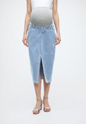 Gravid kvinne iført hvit topp, lyseblå denim graviditetsskjørt med frontsplitt og sølvfargede høyhælte sandaler, stående mot hvit bakgrunn.