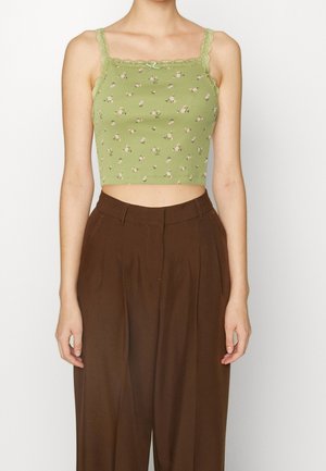 Kobieta w zielonym kwiecistym krótkiem topie z koronkowym wykończeniem i brązowych, szerokich spodniach z wysokim stanem, stojąca na tle białego tła.